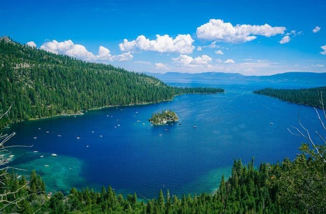 An elevated view of a deep blue bay with a small, forested island, surrounded by pine-covered hills and dotted with boats.