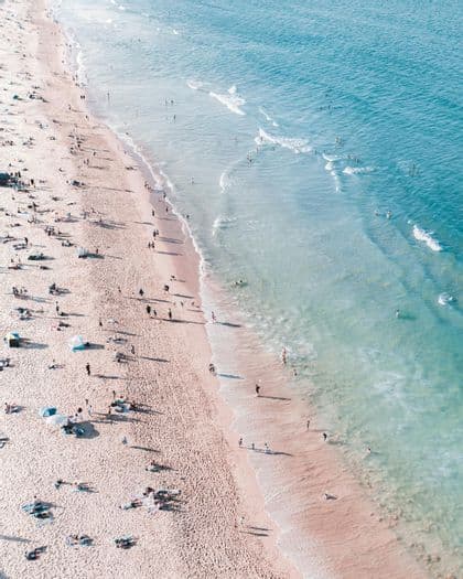 An aerial view of a sandy beach where many people are sunbathing and swimming in the turquoise ocean.