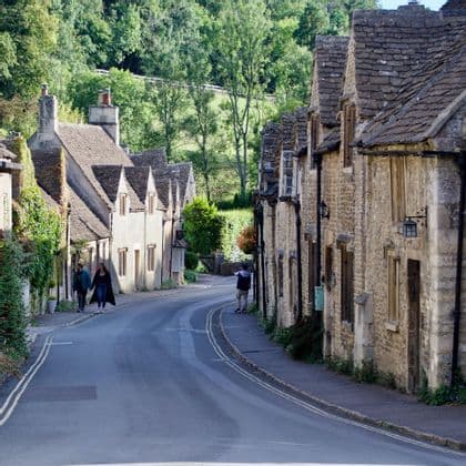 A winding road runs through a village of traditional stone cottages, with people walking on the sidewalks and a green hill in the background.