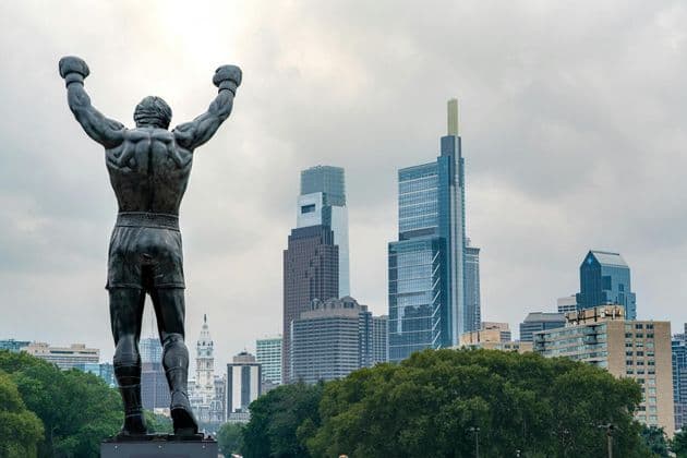 The back of a bronze statue of a boxer with its arms raised in a victory pose, overlooking a city skyline with skyscrapers.