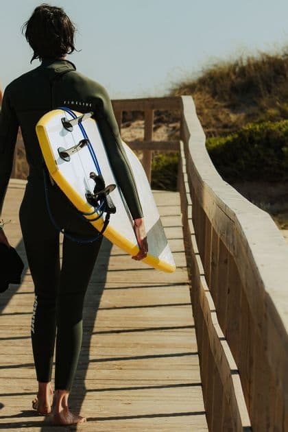 Una persona di spalle in muta cammina a piedi nudi su una passerella di legno portando una tavola da surf.