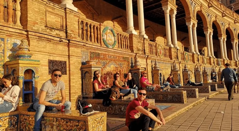Un groupe WeRoad en voyage se détend sur des bancs ornés et carrelés, dans une place ensoleillée aux arcades en briques.