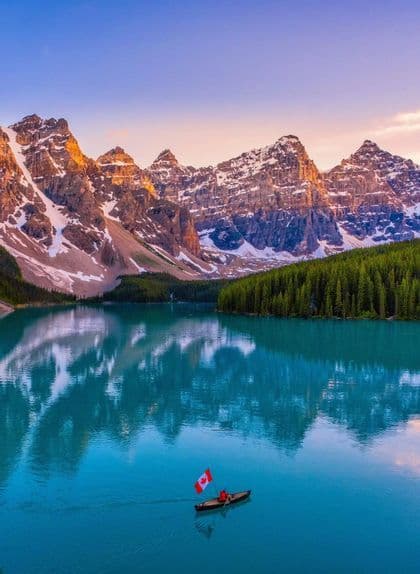 A person in a canoe holding a Canadian flag paddles across a turquoise lake in front of snow-capped mountains and a forest.