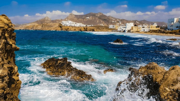 Le onde si infrangono contro una costa rocciosa, con un villaggio costiero di edifici bianchi e montagne sullo sfondo sotto un cielo azzurro.