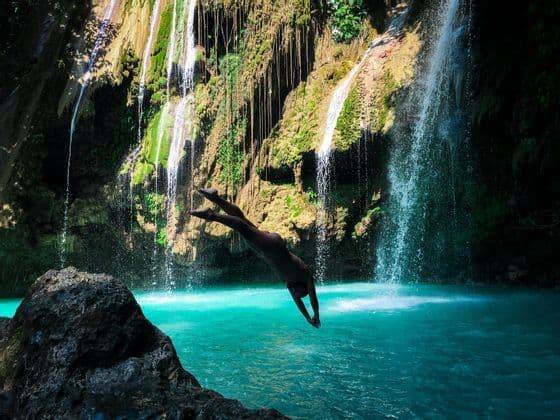 A person dives headfirst from a rock into a turquoise pool at the base of a cascading waterfall.