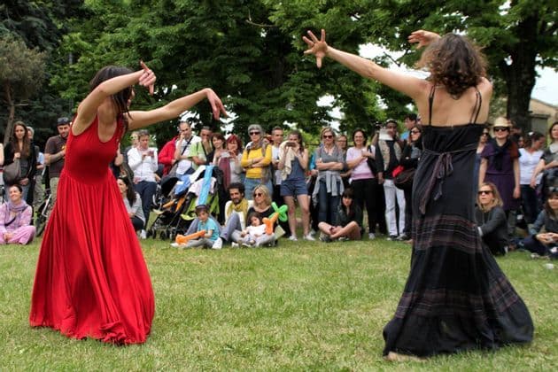 Due donne in abiti lunghi danzano in modo espressivo su un prato verde, mentre una folla osserva in un parco.