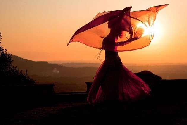 Una donna in silhouette danza con una grande sciarpa traslucida contro un tramonto dorato su colline lontane.