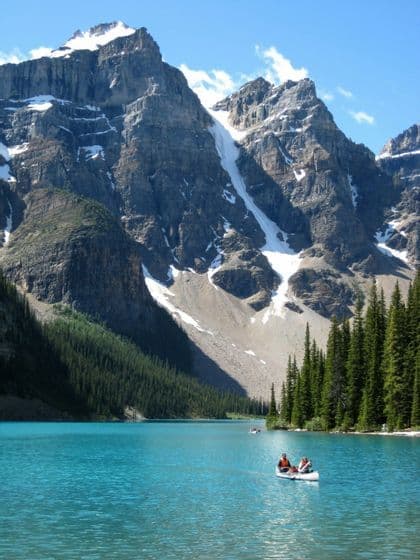A WeRoad group trip paddles a canoe on a bright turquoise lake in front of tall, snow-capped mountains and a pine forest.