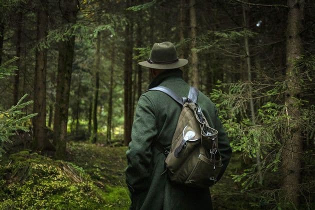 Una persona di spalle, con cappotto verde, cappello e zaino, si trova in una fitta foresta tra alberi alti.