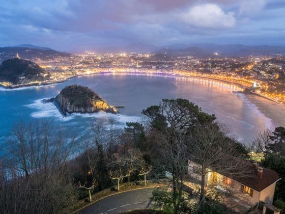 An elevated view of a coastal city at dusk, with lights illuminating a crescent-shaped bay, a beach, and a small island in the water.