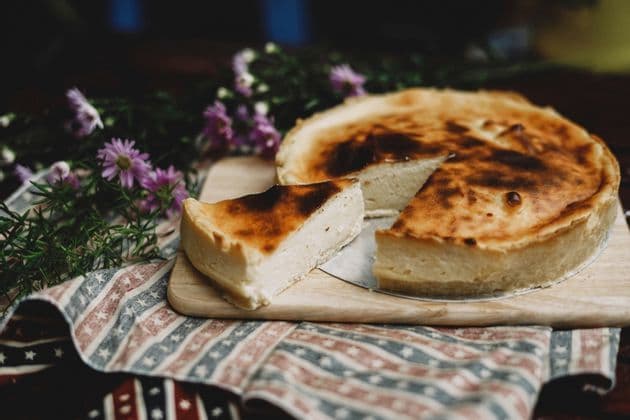 A burnt-top cheesecake with a slice cut out sits on a wooden board, placed on a patterned cloth next to purple flowers.