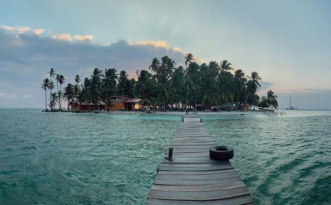 A wooden pier extends over turquoise water towards a small tropical island filled with palm trees and huts at sunset.