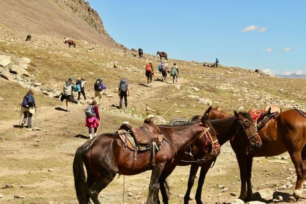 Un groupe WeRoad en randonnée sur un sentier de montagne avec des sacs à dos, des chevaux sellés au premier plan sous un ciel dégagé.