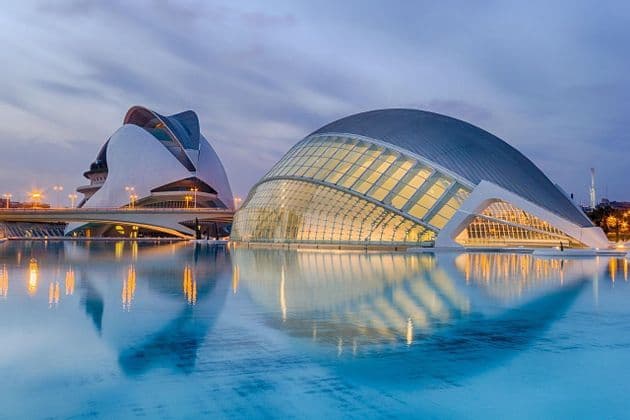 Deux bâtiments modernes et illuminés, aux façades de verre et blanches, se reflètent dans une eau bleue calme au crépuscule.