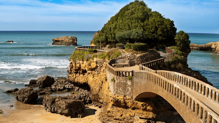 Una passerella in pietra e un ponte ad arco collegano una spiaggia sabbiosa a un isolotto roccioso e alberato, bagnato dall'oceano.