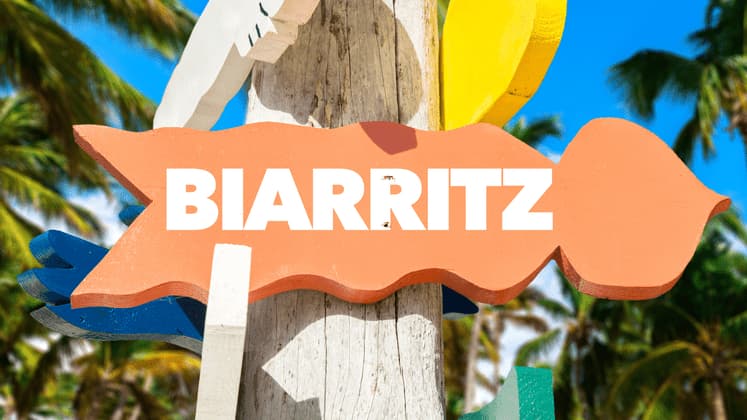Un cartello in legno colorato a forma di pesce con la scritta 'Biarritz', con palme e un cielo blu sullo sfondo.