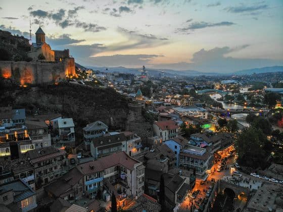 Una veduta dall'alto di una città storica al tramonto, con una fortezza illuminata su una collina che domina case tradizionali e un fiume.