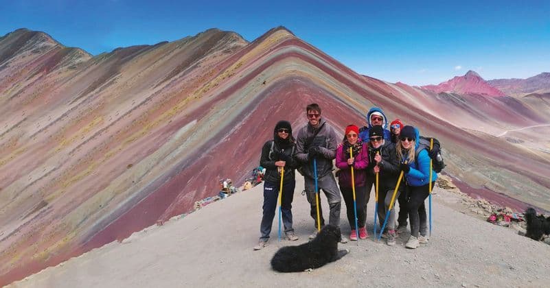Un viaje en grupo de WeRoad posando con bastones de trekking y un perro en una cresta de montaña, con montañas de rayas coloridas de fondo.