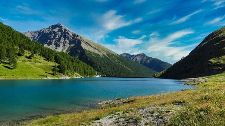 Un lago turchese si trova in una valle tra montagne verdi coperte da foreste di pini, sotto un cielo blu striato di nuvole.