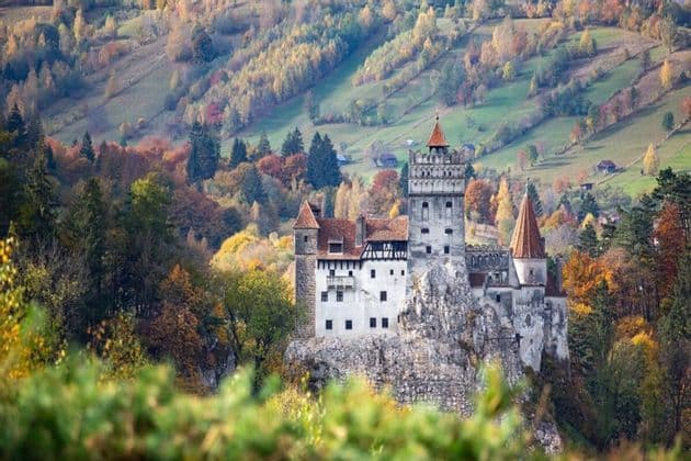 Un castello in pietra con tetti in tegole rosse sorge su uno sperone roccioso, circondato da dolci colline coperte da variopinte foreste autunnali.