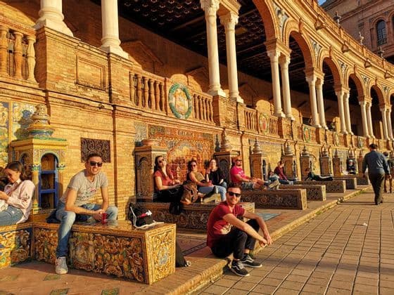 Un voyage de groupe WeRoad se détendant sur des bancs carrelés ornés devant un bâtiment en arcades à l'heure dorée.