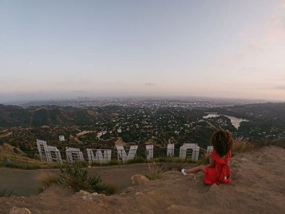 Une femme en robe rouge est assise sur une colline derrière le panneau Hollywood, dominant un vaste paysage urbain au crépuscule.