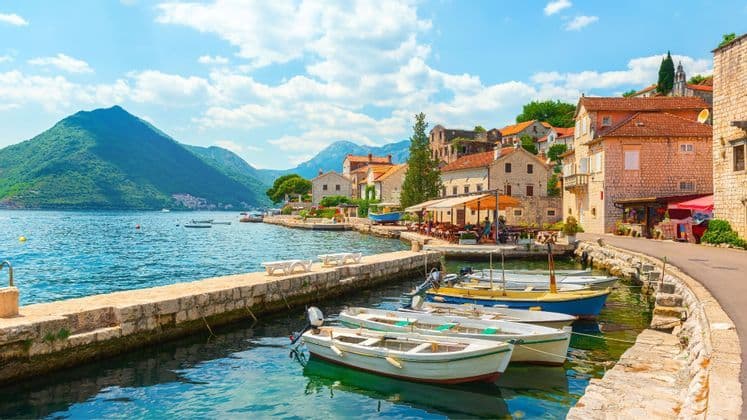 Piccole barche ormeggiate nell'acqua cristallina, accanto a un molo di pietra in un borgo costiero, con montagne verdi sullo sfondo.