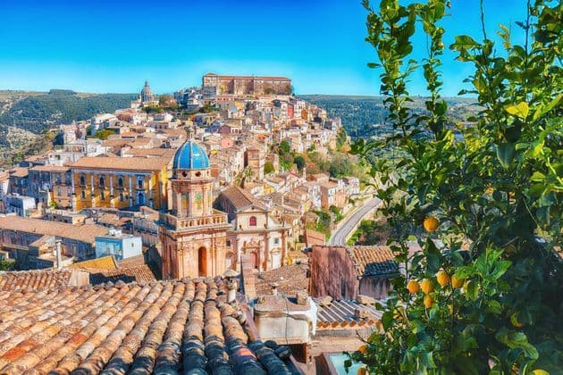 Una veduta soleggiata sui tetti di terracotta di un borgo collinare con una chiesa dalla cupola azzurra, incorniciata da un albero di limoni.