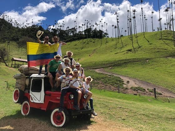 Un groupe WeRoad pose sur une jeep rouge, drapeau colombien en main, dans une vallée verdoyante parsemée de grands palmiers.