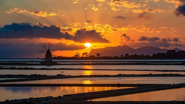 Un mulino a vento tradizionale si staglia contro un tramonto arancione sopra le saline riflettenti, con montagne in lontananza.