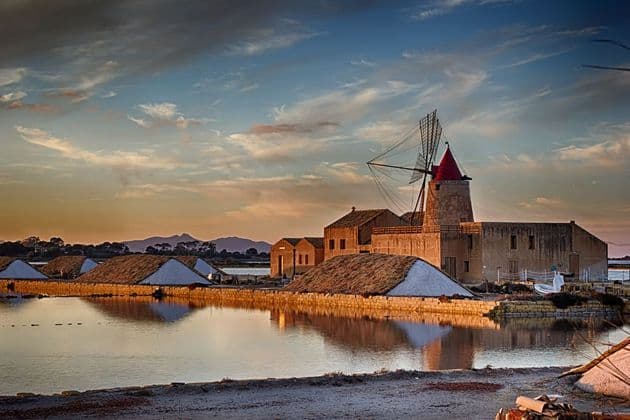 Un mulino a vento tradizionale dal tetto rosso sorge accanto a saline al tramonto, e il suo riflesso è visibile sull'acqua.