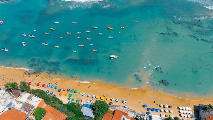 Eine Luftaufnahme eines Sandstrandes mit bunten Sonnenschirmen und Booten, die auf dem türkisfarbenen Meer schwimmen.