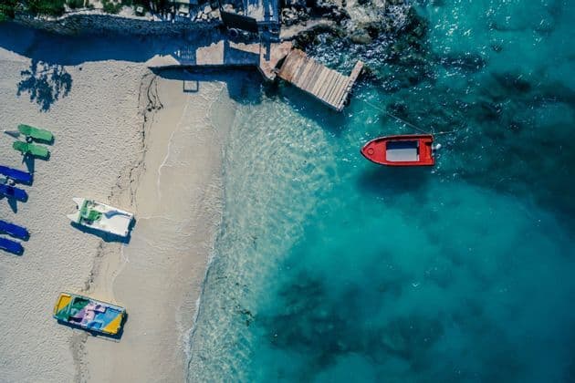 Una vista aérea de una playa de arena con barcos, un pequeño muelle de madera y un barco rojo flotando en agua turquesa clara.