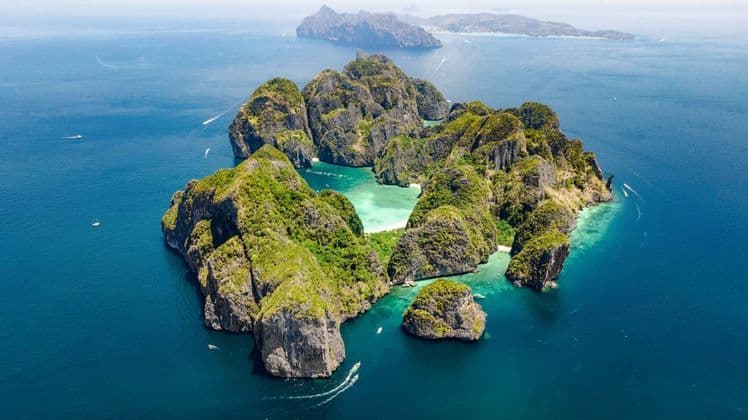 An aerial view of a green, rocky archipelago in a calm blue sea, with small boats sailing around its base.