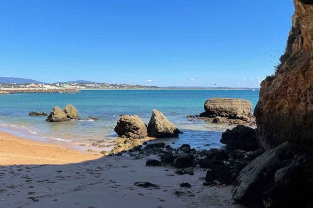 A view from a coastal cave of a sandy beach with large rocks in the turquoise sea under a clear blue sky.