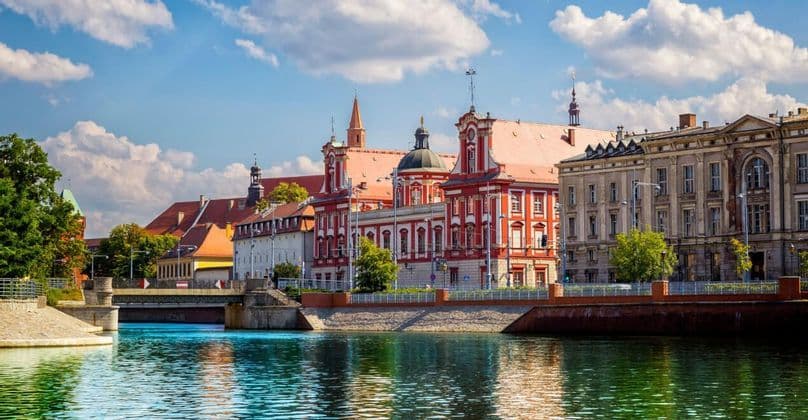 Edifici storici colorati, tra cui uno rosso e bianco in evidenza, costeggiano un fiume in una giornata di sole sotto un cielo nuvoloso.