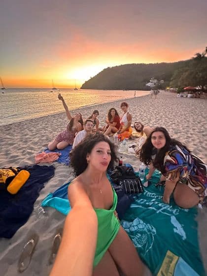 Une femme prend un selfie de son groupe WeRoad lors d'un pique-nique sur une plage de sable, au coucher du soleil coloré.