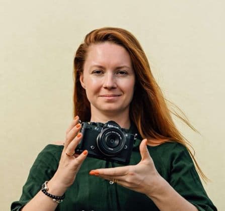 Eine Frau mit langen roten Haaren und einem grünen Oberteil hält eine schwarze Nikon Z spiegellose Kamera mit beiden Händen hoch.
