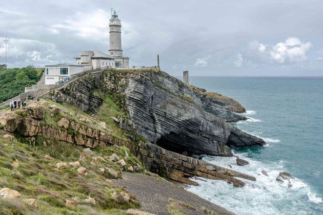 Un gruppo WeRoad percorre un sentiero verso un faro, su una ripida scogliera rocciosa stratificata che domina l'oceano.