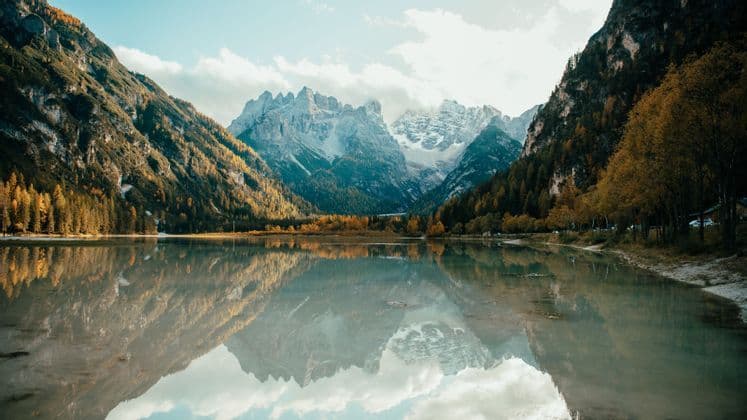 Ein ruhiger See spiegelt eine schneebedeckte Bergkette und einen Wald mit Herbstlaub unter einem teilweise bewölkten Himmel.