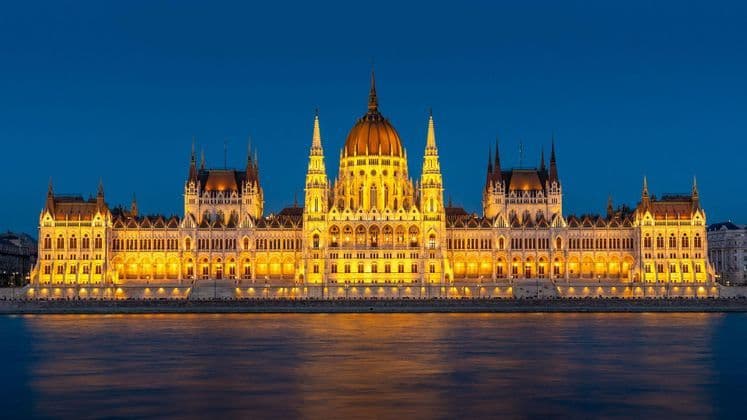 Un grande edificio sontuoso illuminato da luci dorate al crepuscolo, con il suo riflesso visibile sull'acqua sottostante.