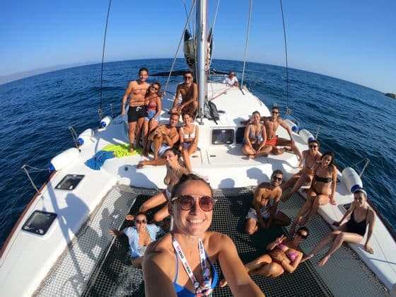 Eine WeRoad-Gruppe lächelt für ein Selfie auf dem Deck und im Netz eines Katamarans auf offener See unter einem klaren blauen Himmel.