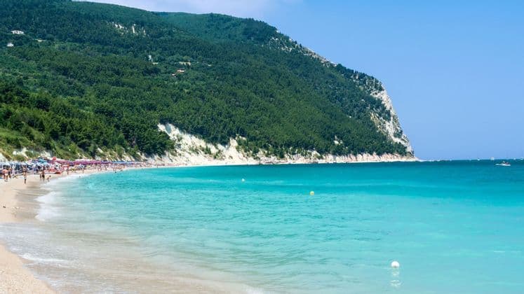 Una spiaggia sabbiosa affollata incontra acque turchesi ai piedi di una grande scogliera alberata sotto un cielo azzurro e limpido.