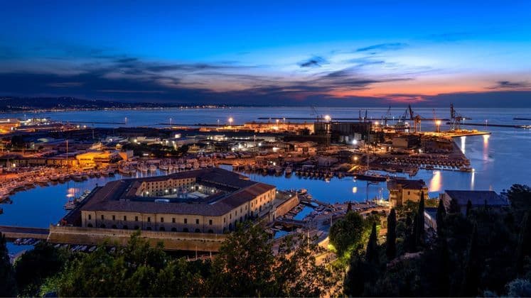 Una città portuale illuminata e il suo porto al tramonto, con un grande edificio storico e barche ormeggiate.