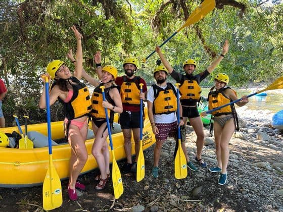 Un viaggio di gruppo WeRoad con caschi e giubbotti di salvataggio in posa con pagaie accanto a un gommone sulla riva di un fiume.