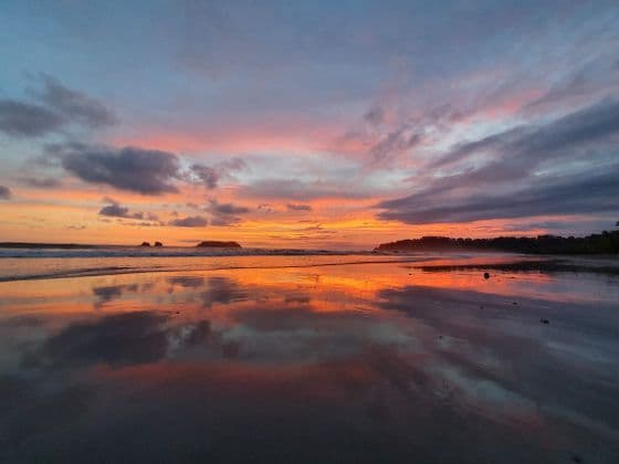 Un tramonto vibrante con nuvole arancioni, rosa e blu che si riflettono sulla sabbia bagnata e specchiata di una spiaggia.