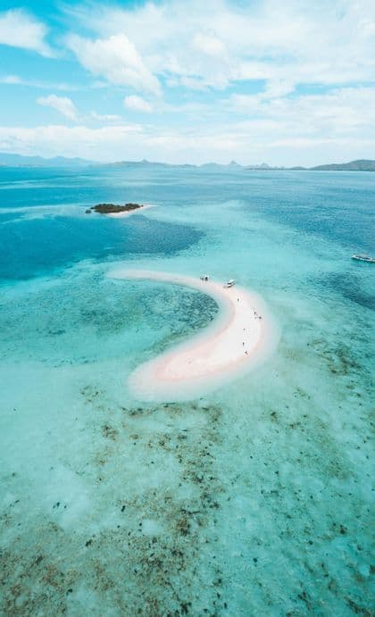 Vue aérienne d'un voyage en groupe WeRoad sur un banc de sable rose en forme de croissant dans des eaux turquoise, avec des bateaux et des îles lointaines.
