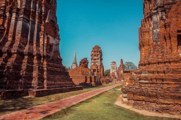 A brick pathway leads through a complex of ancient red brick temple ruins and stupas under a clear blue sky.