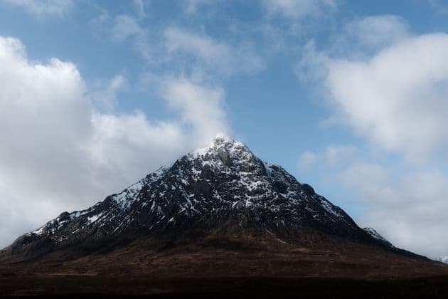 Une montagne rocheuse enneigée se dresse sous un ciel bleu avec des nuages blancs éparpillés.