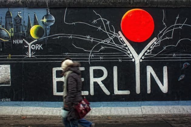Una persona sfocata in movimento passa davanti a un grande murale di graffiti su un muro con la parola 'BERLIN' scritta a grandi lettere bianche.
